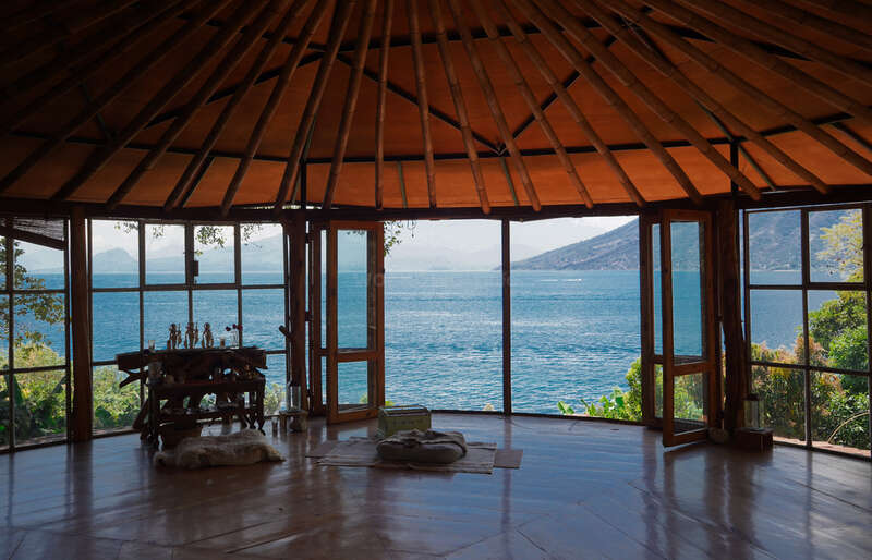 A spacious wooden room with large windows offers a breathtaking view of a tranquil blue lake and distant mountains, creating a serene and peaceful atmosphere inside.