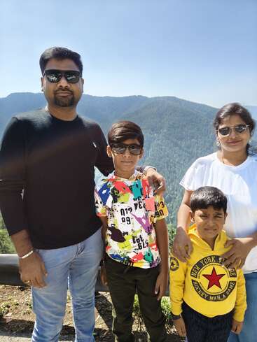 A family of four, all wearing sunglasses, stands outdoors with scenic green mountains and a clear blue sky in the background, posing happily for a photo.
