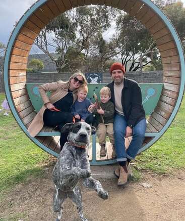 Una familia feliz con dos niños pequeños está sentada dentro de un gran banco circular al aire libre, mientras su enérgico perro blanco y negro fotobombea la imagen en primer plano.