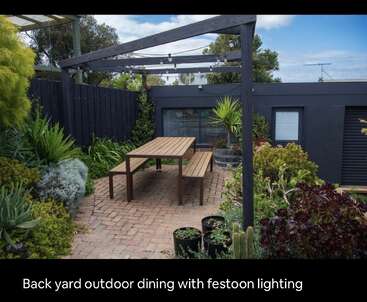 Este acogedor patio trasero cuenta con una mesa de comedor de madera con bancos, rodeada de exuberantes plantas verdes, iluminación de festón, suelo de patio de ladrillo y una valla oscura y moderna.