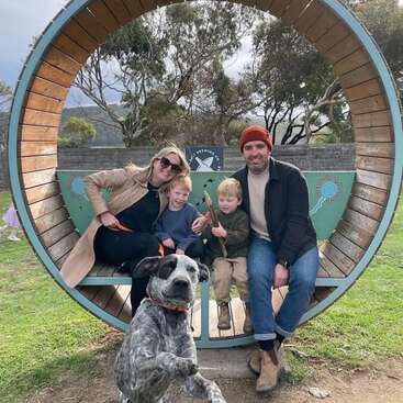 Eine glückliche Familie mit zwei kleinen Jungen sitzt auf einer großen, runden Bank im Freien, während ihr energiegeladener schwarz-weißer Hund im Vordergrund das Bild förmlich bombardiert.