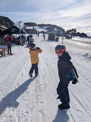 Two children in winter gear walk on a snowy path near a ski lodge. People ski, relax outside, and cars are parked nearby. Mountains and trees surround.