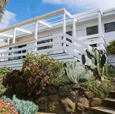 A modern white house with a large deck and glass railings is surrounded by a lush desert garden, featuring succulents, cacti, rocks, and blue sky above.