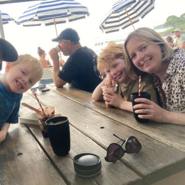 Eine lächelnde Frau und zwei Kinder sitzen an einem Holztisch im Freien und genießen Getränke mit gestreiften Strohhalmen. Sonnenschirme beschatten die Umgebung, Sonnenbrillen liegen auf dem Tisch.