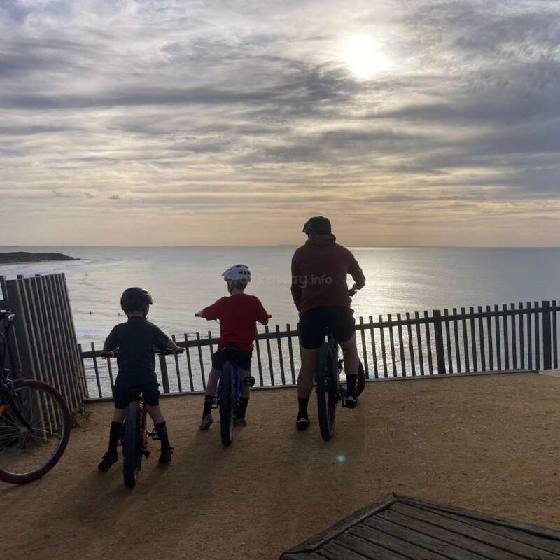 Tres personas en bicicleta, con casco, contemplan el océano desde un mirador junto a un acantilado al atardecer. La escena es apacible, con aguas tranquilas y nubes espectaculares.
