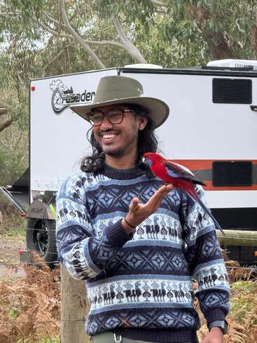 Um homem sorridente com um suéter estampado e chapéu está ao ar livre, segurando um papagaio vermelho vibrante na mão. Uma van e árvores estão atrás dele.