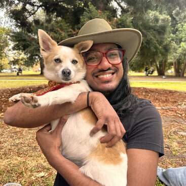 A person wearing a hat and red glasses smiles while hugging a fluffy dog in a park. The scene feels joyful, warm, and surrounded by nature.