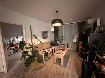 Uma sala de estar aconchegante com um sofá bege, manta listrada, várias plantas, mesa de centro de madeira, estante, quadro e iluminação suave. Você pode ver um cômodo adjacente.