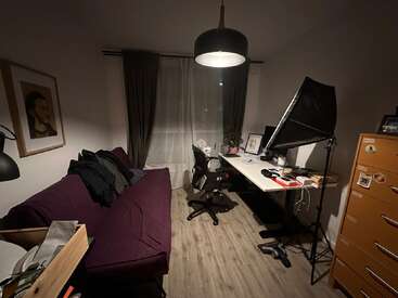Um home office aconchegante e pouco iluminado apresenta um sofá roxo, mesa de trabalho com computador, cadeira de escritório, luz softbox, gavetas laranja, janela grande e obras de arte emolduradas.