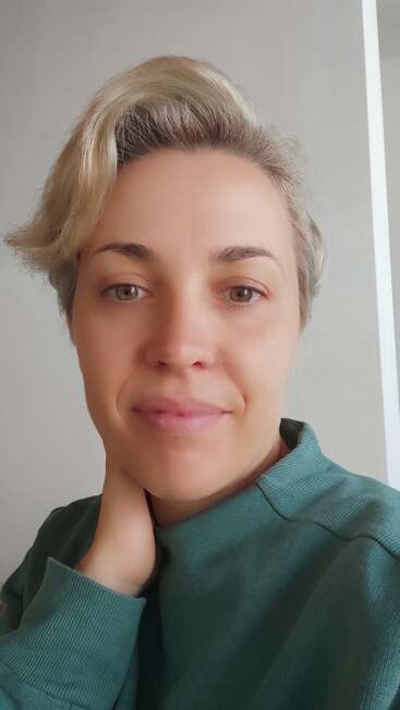 A imagem mostra uma mulher com cabelo loiro curto, vestindo um suéter verde, posando para uma selfie em frente a uma parede branca com uma linha branca vertical no lado direito.