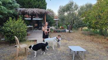 A woman holds a child, while another child stands beside two dogs in a rustic garden with trees, a wooden shelter, chairs, and a table outside.