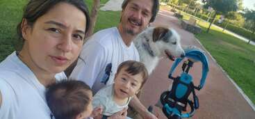 A family sits on a park bench: mother holding a baby, father holding a toddler, a stroller, and a happy dog beside them, enjoying outdoors.
