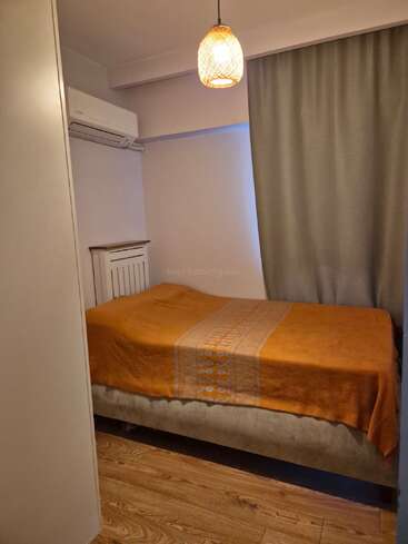A small, cozy bedroom features a single bed with an orange blanket, wooden floor, grey curtains, pendant light, air conditioner, and minimal, clean decor.