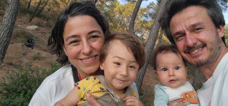 A smiling family of four poses closely together outdoors among tall trees. The parents hold two young children, radiating happiness and warmth during a nature outing.
