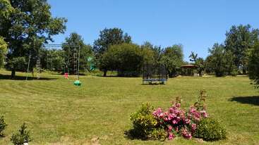 A sunny park scene with green grass, trees, a swing set, and trampoline. Pink flowers bloom in the foreground, and a gazebo appears in the background.