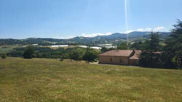 A small house with a red-tiled roof sits in a grassy field. Rolling hills, trees, greenhouses, and mountains stretch out beneath a clear blue sky.