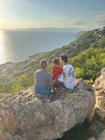 A family of three sits on a large rock, overlooking a scenic coastal view with sparkling water, green hills, sunset sky, and distant mountains. Peaceful moment.
