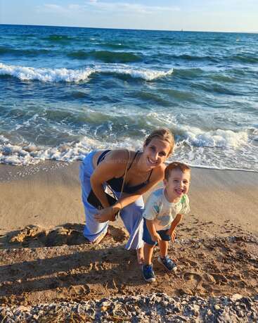 A smiling woman and a young boy happily pose together on a sandy beach, with gentle waves behind them under a bright, clear blue sky.