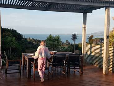 Una mujer se encuentra de pie en una terraza de madera con muebles de exterior, contemplando la exuberante vegetación y las vistas del océano azul bajo una pérgola parcialmente sombreada, mientras disfruta del paisaje.
