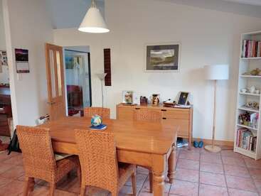 Esta imagen muestra un acogedor comedor con una mesa de madera, cuatro sillas de mimbre, un aparador, estanterías, lámparas de pie, obras de arte y objetos decorativos expuestos.