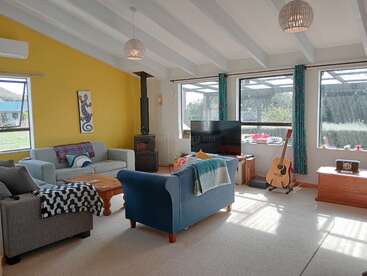 Este acogedor salón cuenta con paredes de color amarillo brillante, sofás modernos, una estufa de leña, grandes ventanales, un televisor, una guitarra y una decoración alegre y acogedora, con mucha luz natural.