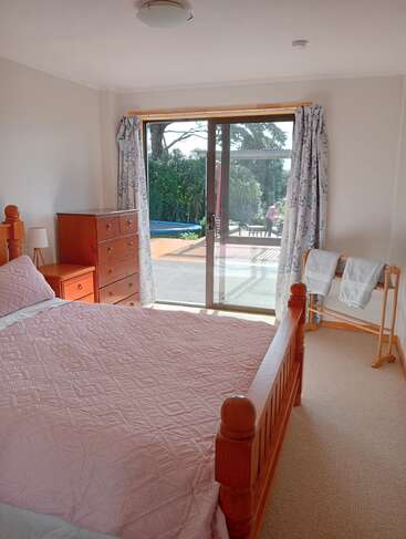 Un acogedor dormitorio con una cama de madera, una colcha rosa, una cómoda, una lámpara y un toallero. Las puertas correderas de cristal dan a un soleado patio exterior.