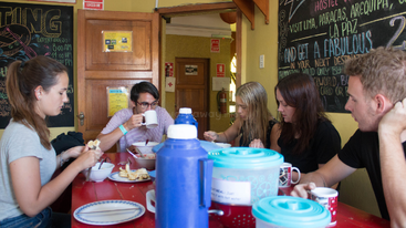 Five young adults sit around a red table eating breakfast together. Blue thermoses, bowls, and mugs are present. Chalkboard walls advertise hostel tours and destinations.