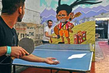 Das Bild zeigt zwei Männer, die Tischtennis spielen, wobei der Arm des einen Mannes zu sehen ist, der einen Schläger hält und der andere Mann im Hintergrund sich auf den Aufschlag vorbereitet.