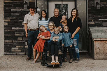 A multigenerational family stands smiling outside a house. Adults and children gather around a seated elderly woman in a wheelchair, exuding warmth and togetherness.