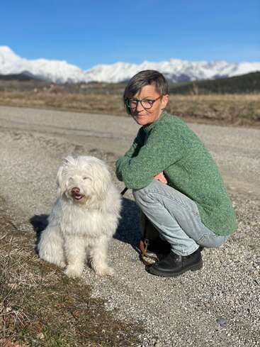 Uma pessoa de cabelo curto e óculos está agachada ao lado de um cachorro branco e fofo em um caminho de cascalho, com montanhas cobertas de neve e um céu azul claro ao fundo.