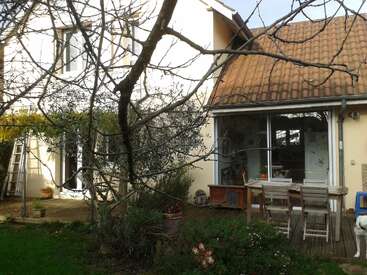 A quaint house with a tiled roof, glass doors, and a wooden patio set. Leafless tree in foreground, ladder, garden plants, and partial dog visible.