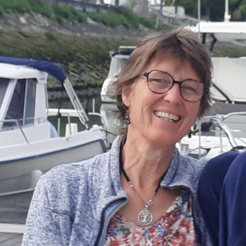 A cheerful woman with short hair and glasses smiles by a marina. She wears a floral top, blue jacket, and necklace. Boats and water are visible behind her.