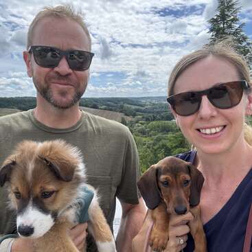 A smiling couple wearing sunglasses holds two adorable puppies outdoors. Behind them, lush green hills and a cloudy blue sky provide a beautiful scenic backdrop.