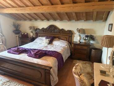 Este acogedor dormitorio cuenta con una gran cama de madera ornamentada, mesillas de noche vintage, decoración floral, detalles en morado, un techo inclinado de madera, lámparas y un ambiente acogedor y rústico.