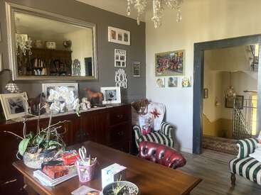 Un acogedor salón con sillones a rayas, un escritorio de madera, fotos enmarcadas, un gran espejo de pared, plantas de interior, lámparas de araña y un pasillo con escaleras a la vista.