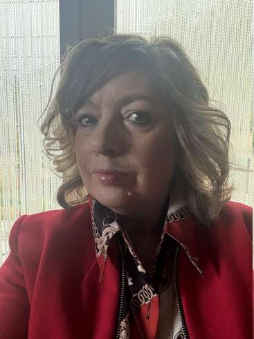 Una mujer de pelo ondulado y ojos expresivos lleva una chaqueta roja y un pañuelo estampado, y posa en un interior frente a una ventana con cortinas transparentes.