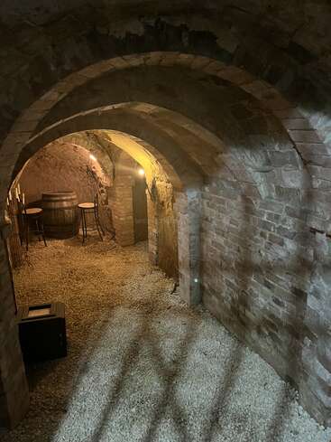 Esta imagen muestra una antigua bodega de piedra con techos arqueados, suelo de grava, iluminación tenue, un barril de vino, mesas pequeñas y sillas en un ambiente rústico.