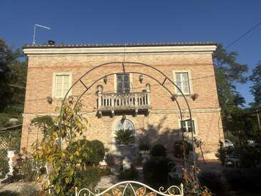 Una encantadora casa de ladrillo de dos plantas con ventanas arqueadas, un balcón central, arbustos bien recortados y un arco en el jardín, todo bañado por la luz dorada del sol.