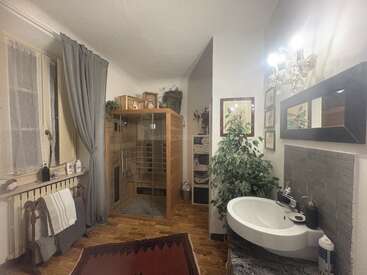 Este cuarto de baño cuenta con una sauna de madera, un lavabo blanco, un elegante espejo, una planta en maceta, una acogedora ventana con cortinas, toallas, cuadros enmarcados y una alfombra estampada. Iluminación cálida.