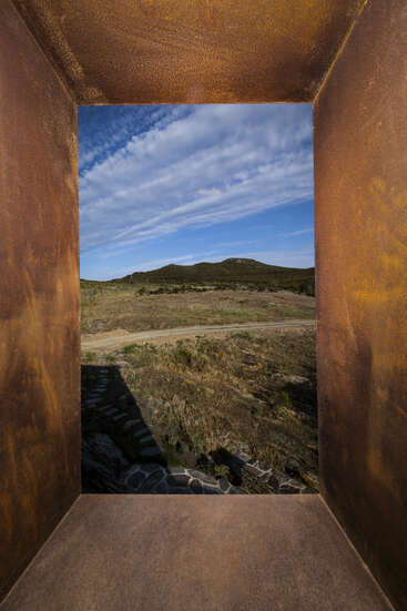 A rust-colored, rectangular metal frame perfectly outlines a scenic landscape of hills, clouds, and blue sky, creating a natural picture window effect in nature.