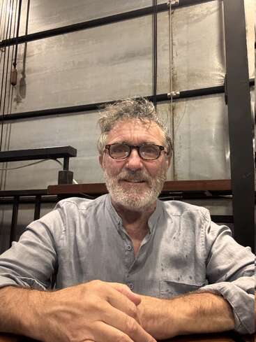 A bearded man with glasses sits at a table, smiling. He wears a light grey shirt. The industrial background features metal beams and a concrete wall.