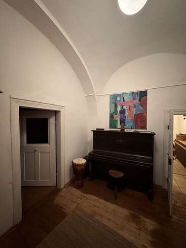 A warmly lit room with arched ceilings features a black upright piano, a drum, a stool, abstract wall art, wooden floors, and two open doorways.