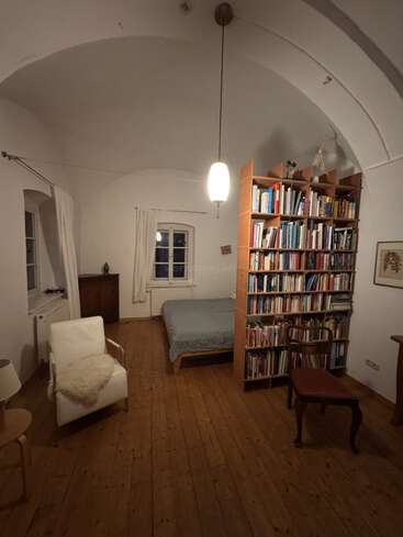 A cozy, softly lit room features a bed, tall bookshelf, armchair, wooden chair, and table on wooden floors, with white walls and arched ceiling.