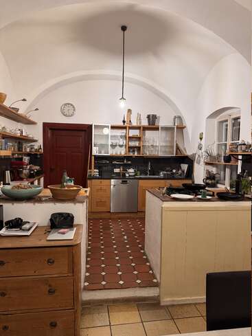 This image shows a cozy kitchen with arched ceilings, wooden cabinets, open shelves, a red door, tiled floors, various utensils, and a warm, inviting atmosphere.