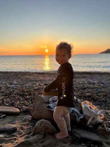 A young child smiles while standing barefoot on a rocky beach at sunset, with warm sunlight reflecting on the water and a picturesque, serene atmosphere.