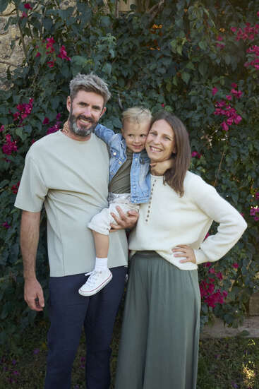 A family of three poses happily outdoors, standing in front of green foliage with vibrant pink flowers, smiling warmly and embracing each other on a sunny day.