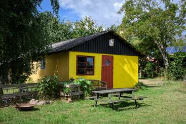 The image shows a small, wooden building with a sloping roof, featuring a large window and a door, situated in a grassy area with trees and a body of water in the background.