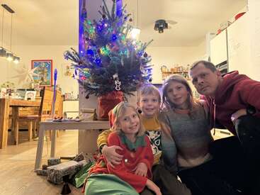 A family of four sits together by a decorated Christmas tree in a cozy, warmly lit living room, smiling and celebrating the holiday season with gifts.