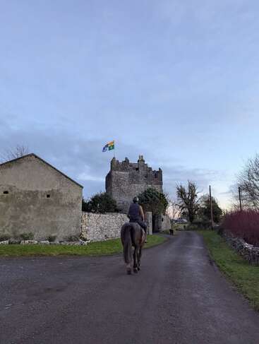 Une personne chevauche un cheval sur une route de campagne en direction d'un château en pierre surmonté d'un drapeau. De vieux bâtiments, des arbres dénudés et un ciel nuageux complètent le tableau.