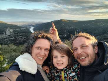Une famille souriante de trois personnes prend un selfie en plein air avec des collines verdoyantes, une rivière et des nuages spectaculaires en arrière-plan. Une personne montre du doigt la vue.